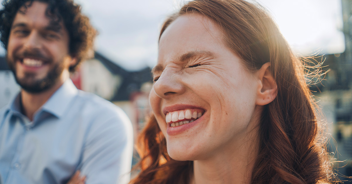 Rothaarige Frau und Mann, der älter ist als sie mit braunen Locken lachen.