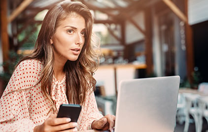 Frau mit langen, brauen Haaren sitzt am Laptop und hält Handy in der Hand