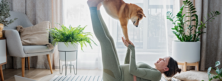 Frau liegt auf dem Rücken und macht Gymnastik. Dabei balanciert sie ihren Hund auf Händen und Füßen. 