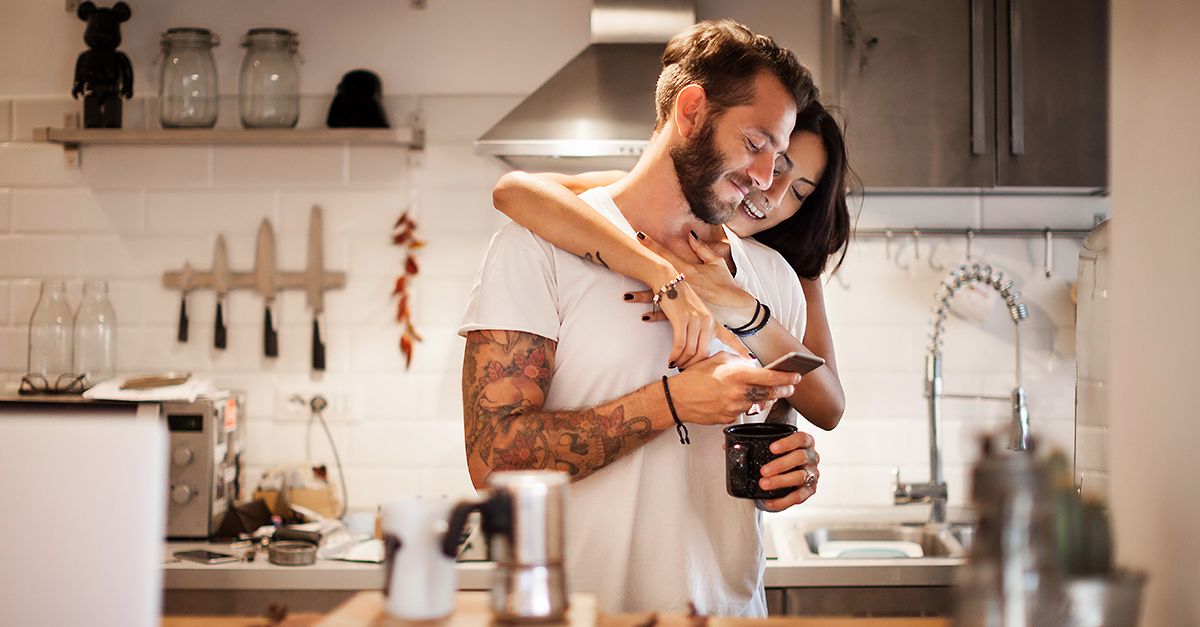 Frau umarmt Mann in Küche, beide blicken grinsend auf Smartphone