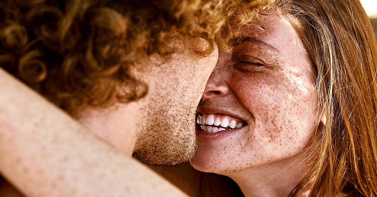 Rothaarige Frau mit Sommersprossen umarmt Mann mit Locken, die Gesichter dicht beieinander. Sie lächelt.