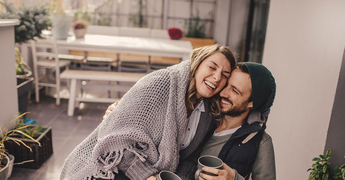 Mann mit Mütze und Frau mit einer Wolldecke im den Schultern lehnen aneinander und lachen.