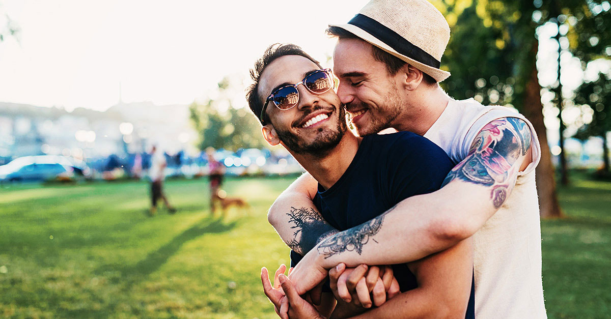 Mann mit Hut und Tattoos umarmt Mann mit Sonnenbrille, beide lachen. 