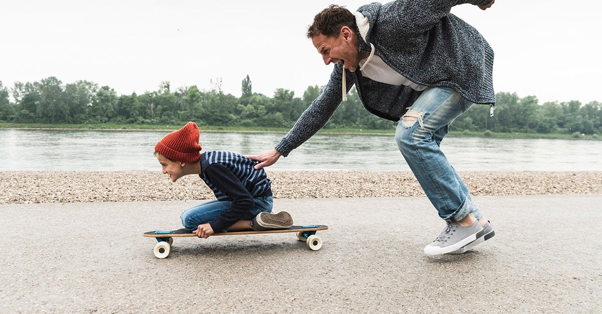 Kind sitzt auf Skateboard, Mann schiebt Kind an, beide lachen. 