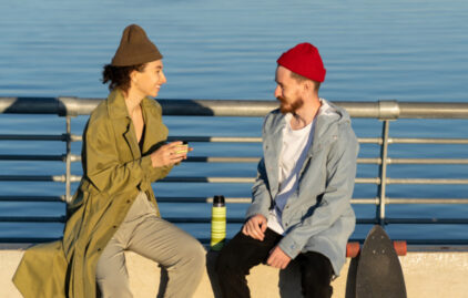 Eine Frau mit grüner Tunika und braunen Haaren und ein Mann mit roter Mütze sitzen an einer Promenade am Meer und unterhalten sich