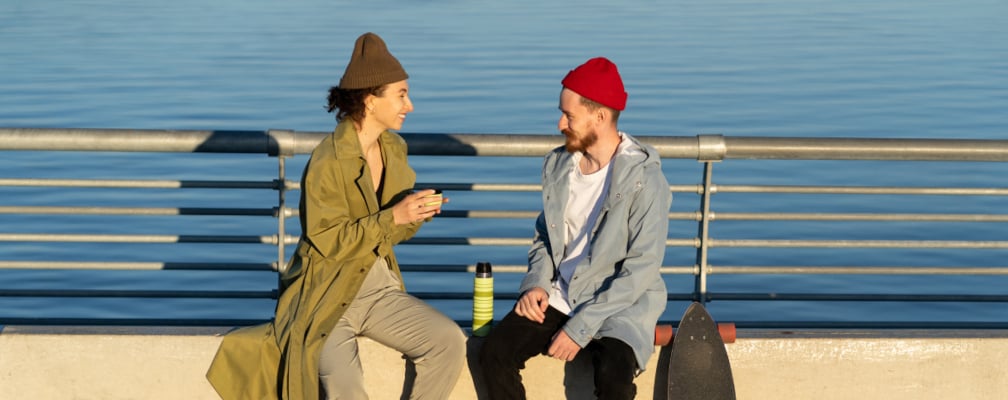 Eine Frau mit grüner Tunika und braunen Haaren und ein Mann mit roter Mütze sitzen an einer Promenade am Meer und unterhalten sich