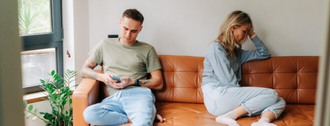 Ein junges Paar sitzt auf einer Couch. Während der Mann am Handy ist, sitzt die Frau abgewandt ein Stück neben ihm. Dei Stimmung wirkt angespannt.