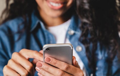 Im Vordergrund wird ein Handy gehalten, im Hintergrund ist eine lächelnde Frau zu erkennen.