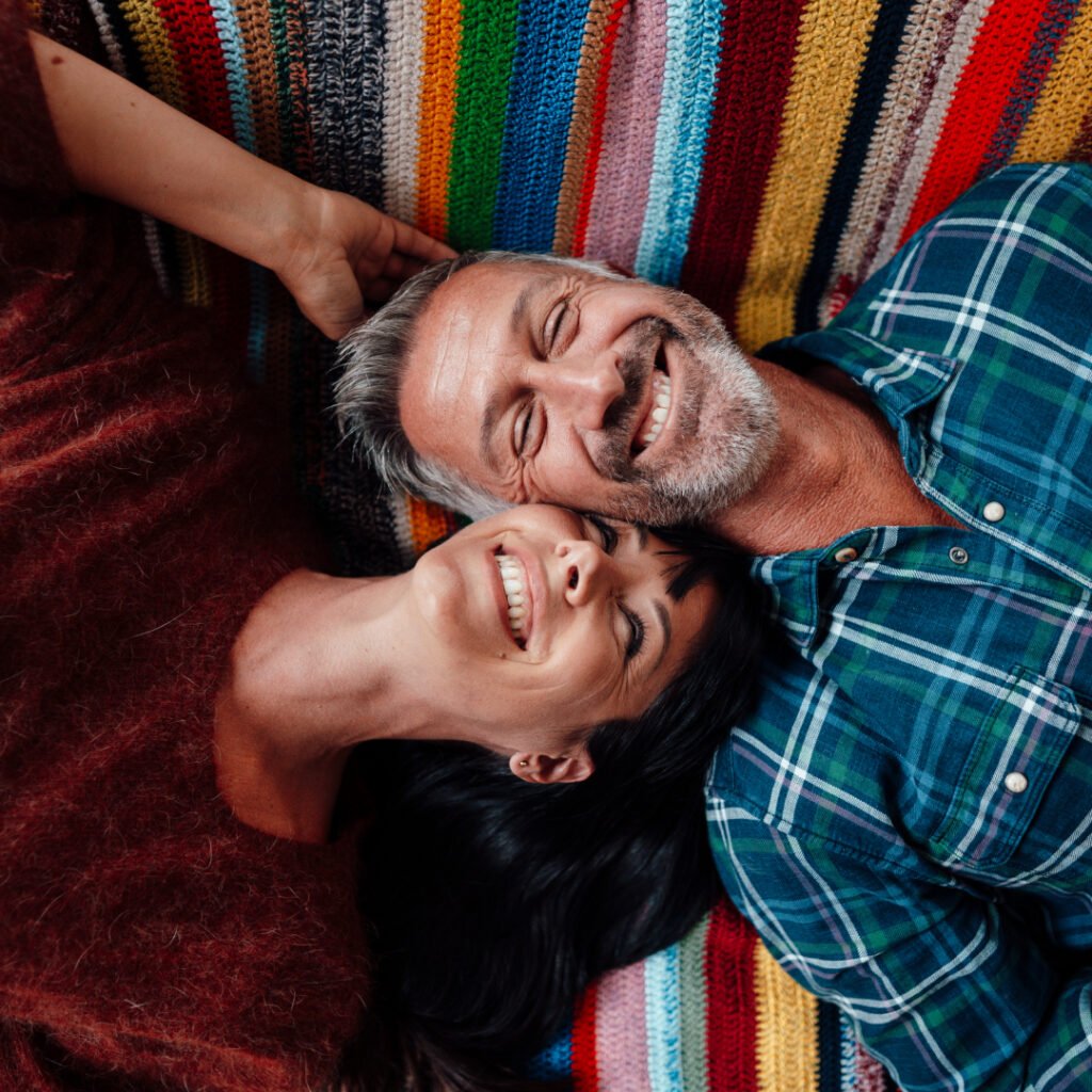 Ein Mann mit grau meliertem Bart und eine brünette Frau liegen rücklings auf einer Couch, lehnen die Köpfe aneinander und lächeln