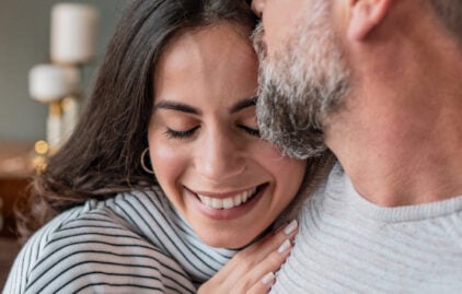 Eine Frau mit schwarzem Haar lehnt sich lächeln an die Schulter eines Mannes mit grau meliertem Bart